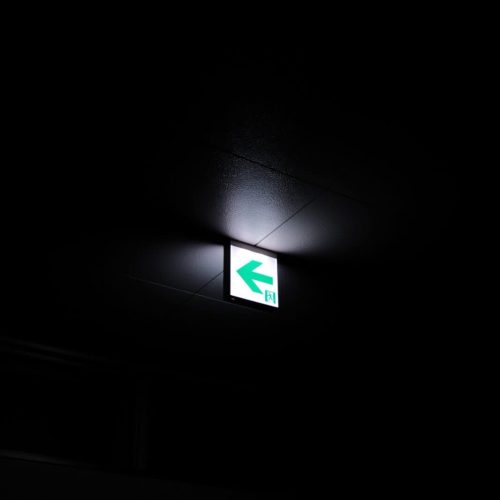 Vertical grayscale selective focus shot of a lighted green exit sign on a ceiling